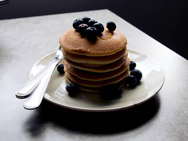 Pancakes with blueberries, stacked on a white plate with a knife and fork. Placed on a grey tabletop.