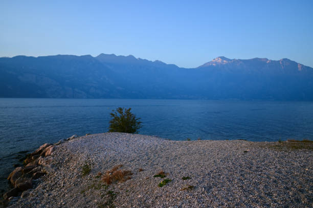 Calm water of garda lake with pebble beach and monte baldo mountain range in background at dusk