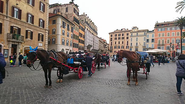Rome, Italy - 4.3.2018: Horse-drawn carriages wait in the bustling Piazza di Spagna near Spanish Steps surrounded by historical buildings and tourists exploring the area under a cloudy sky