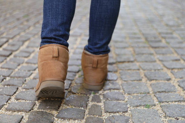 A person is walking on a cobblestone street, with a focus on their legs and footwear. They are wearing slim-fitting dark blue jeans and light brown suede boots.