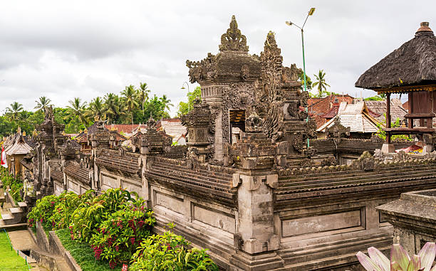 istock Ornate stone temple wall in Bali with intricate carvings. Lush greenery and distant rooftops under a cloudy sky. 2206412088