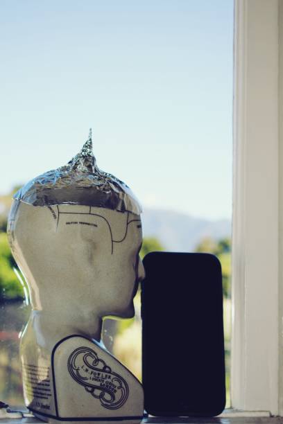 A Phrenology head wearing a tin foil hat looking out of a window with a mobile phone device. This is for a conspiracy and communication conceptual image of either information or disinformation.