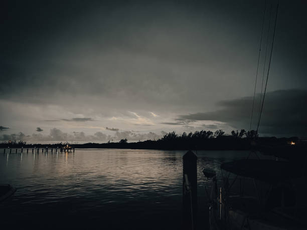 Dark clouds gather over a calm body of water, reflecting the dim light of dusk. A wooden pier extends into the water, while trees line the shore, adding depth to the scene.