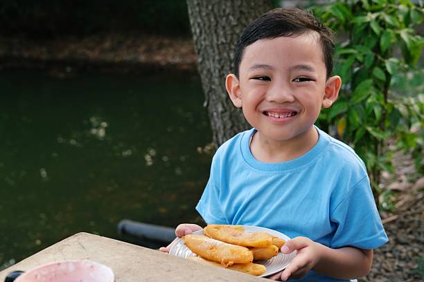 Smiling young boy holding a plate of fried snacks by the riverside. Perfect for food, childhood joy, outdoor lifestyle, family moments, and cultural or traditional meal themes.