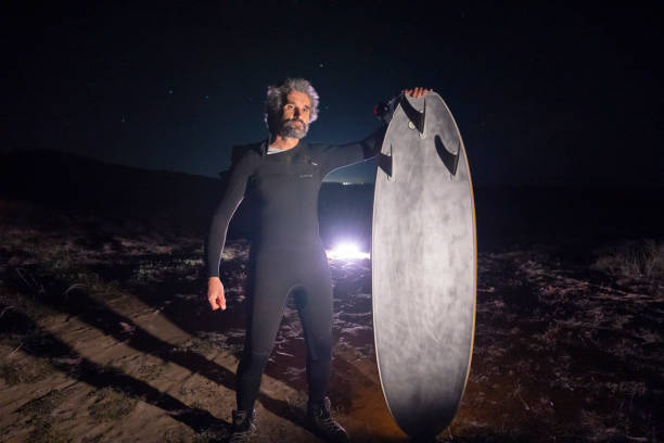 Confident mature surfer proudly displays his surfboard under the dark night sky, illuminated by nearby car headlights, expressing joy and achievement.