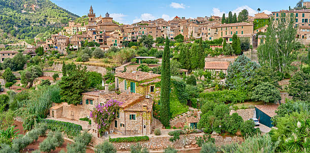 Scenic view of a picturesque village in Majorca, Spain nestled among vibrant green trees and rolling hills. The Mediterranean architecture beautifully complements the serenity of the natural landscape.