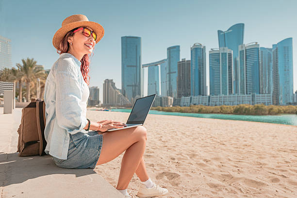 istock Young woman using laptop while sitting on sandy beach with Abu Dhabi skyline in background 2206439088