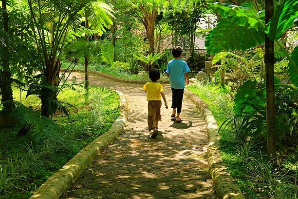 Two children walking on a winding stone path through a lush green tropical garden, surrounded by tall plants and trees, enjoying a peaceful outdoor adventure together.