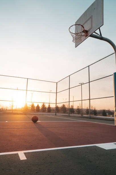 A basketball rests on the court as the sun sets behind the hoop.