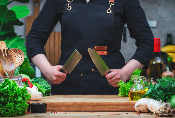 Chef in black holds two sharp Japanese knives in hands. Cozy kitchen with wooden table, kitchenware, vegetables, herbs and ingredients for cooking. Healthy vegan eating, culinary, recipes, food blogging el chef de negro sostiene dos afilados cuchillos japoneses en las manos. acogedora cocina con mesa de madera, menaje de cocina, verduras, hierbas e ingredientes para cocinar. alimentación vegana saludable, culinaria, recetas, blogs de comida