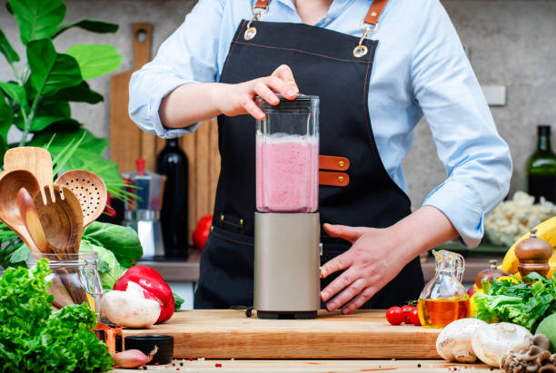 istock Chef makes banana and raspberry smoothie in blender. Cozy kitchen with wooden table, kitchenware, vegetables, herbs and ingredients for cooking. Healthy vegan eating, culinary, recipes, blogging 2206458174