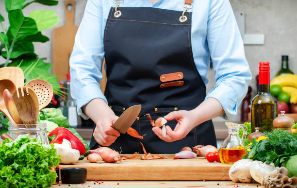 istock Cook peels shallots onion with chef knife. Cozy kitchen with wooden table, kitchenware, vegetables, herbs and ingredients for cooking. Healthy vegan food, culinary, recipes, blogging 2206458193