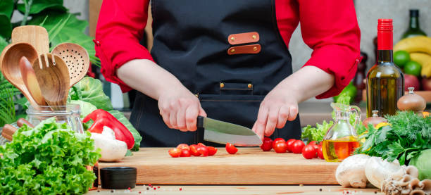 istock Chef cuts cherry tomatoes for salad. Cozy kitchen with wooden table, kitchenware, vegetables, herbs and ingredients for cooking. Home food, cooking, recipes, blogging banner 2206458197
