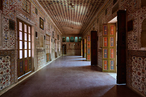 A dynamic, low-angle shot focusing on the rooftop architecture of a historic palace in Rajasthan, India. The structure is built from warm, red/terracotta sandstone and features a row of domed Chatri pavilions, intricate Jaali screen windows, and a crenellated battlement parapet. The details are sharply contrasted against a clear, bright blue sky, emphasizing the traditional Rajput design and craftsmanship.