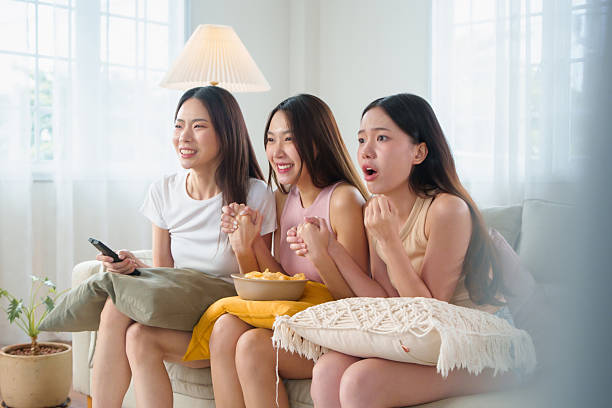 istock Three Asian friends sit closely together on a couch, reacting with excitement while watching at Tv and sharing snacks in a Living room. 2206471736