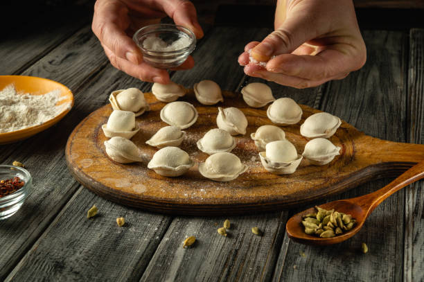 istock Hands carefully sprinkle salt over freshly made dumplings arranged on a wooden board. Various spices and flour are nearby in a cozy kitchen atmosphere 2206488910
