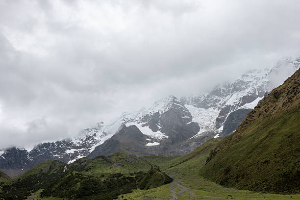 Clouds envelop the snowy mountain peaks, creating a misty, serene landscape. Lush green foothills contrast the rugged terrain.