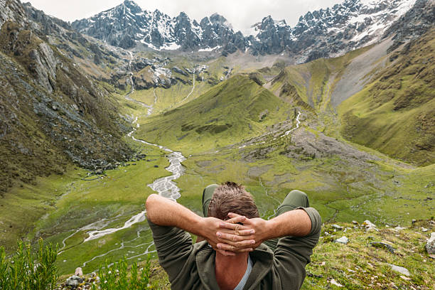 Man sitting on rock enjoying panoramic view of lush mountain valley. Hiker resting with breathtaking landscape in the background. Nature enthusiast appreciating tranquil scenery.