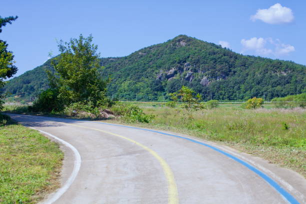 Naju City, South Korea - September 24, 2020: A smooth cycling path winds through the rural landscape along the Yeongsangang Bike Path, with a forested mountain towering in the background under clear blue skies.
