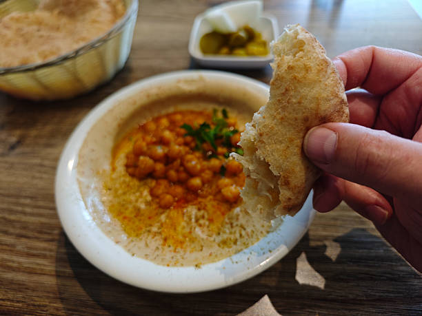 A close-up of a hand holding a piece of fresh pita bread, ready to scoop into a bowl of creamy hummus topped with seasoned chickpeas, olive oil, and parsley. The background includes a basket of pita and a small dish of pickled vegetables, capturing the essence of Middle Eastern cuisine in a cozy dining setting.