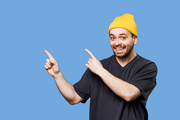 istock Joyful man in yellow beanie points both ways against blue background 2206515996