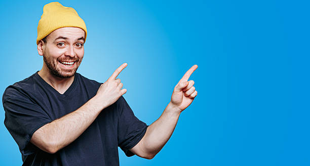 istock Happy man in yellow beanie points in excitement against blue backdrop 2206516006