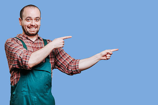 istock Smiling man in overalls pointing to the side against a blue background 2206518069