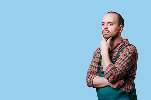 istock Man in plaid shirt and green apron deep in thought against light backdrop 2206518081
