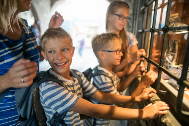 A mother and her three children are gathered around a barred window, looking inside a shop display. The two young boys in matching striped shirts and backpacks are leaning forward with curiosity, while one of them turns his head with a bright smile. Arched passage in historic town of Sirmione.\nShot with Nikon D850