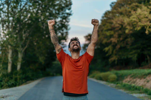 Sportsman celebrating victory raising arms on a road Young sportsman celebrating victory with arms raised, running along a road surrounded by a lush forest, radiating joy and determination arm tattoo ideas for men pictures stock pictures, royalty-free photos & images