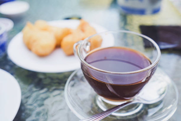 A clear glass cup of hot coffee sits on a saucer with a spoon, accompanied by a plate of pastries on a textured glass table. The scene suggests a relaxing morning or afternoon break.