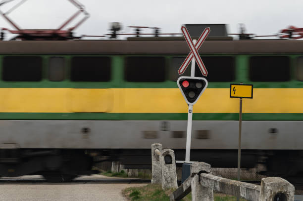 A green and yellow electric locomotive speeds through a railroad crossing, with the red warning signal illuminated. The motion blur of the train emphasizes its speed, while the crossing barriers and signs highlight safety precautions.