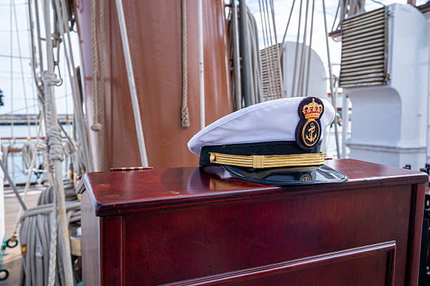 A white hat with a crown on it sits on a wooden table. The hat is a sailor's hat and is placed on a table that is near a large ship. The hat is a symbol of authority and leadership