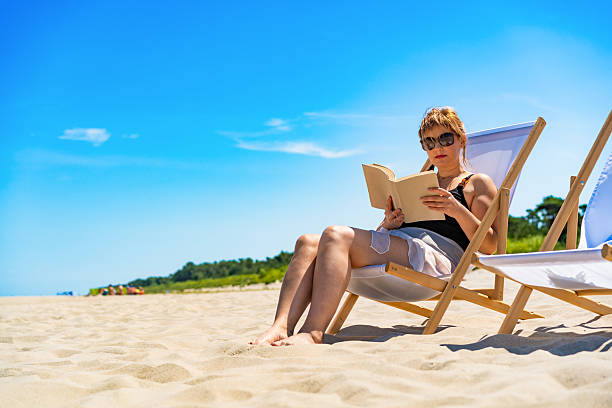 istock Mature women reading books sitting on deckchairs on beach 2206535513