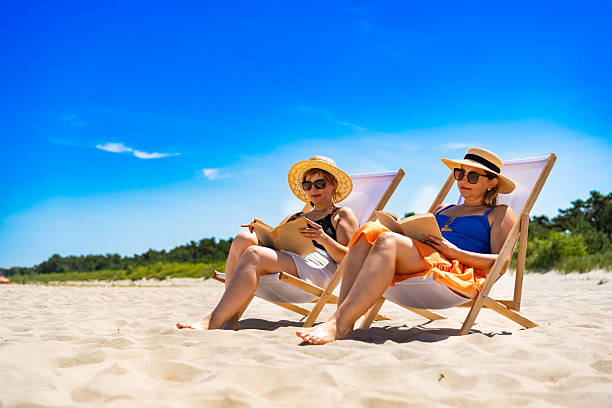 istock Two mature women reading books sitting on deckchairs on beach 2206535530
