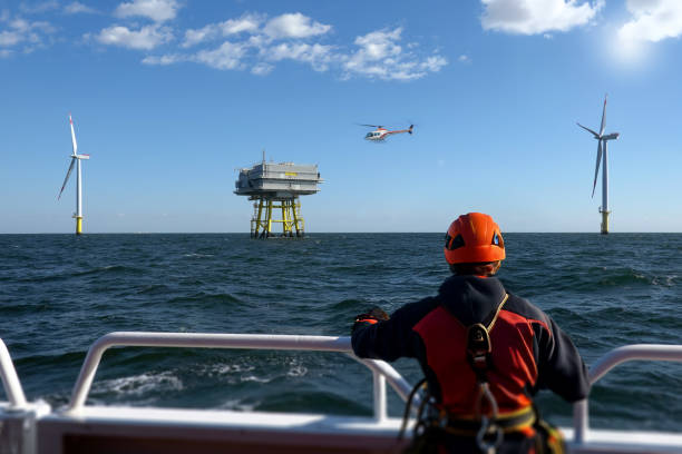 A worker in safety gear on the deck of a boat oversees an offshore wind farm. In the background, two wind turbines and a service platform can be seen. A helicopter flies in the sky, providing transportation to the turbines. The photograph captures the challenging working conditions in the energy sector and the efforts towards sustainability.
