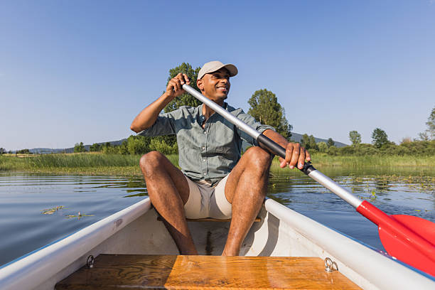 Young man laughing and having fun while canoeing on the calm lake surrounded by stunning nature, front view. Riverside leisure concept.