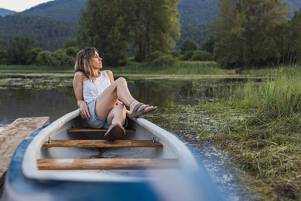 A woman in her 20s paddles a canoe on a serene lake with lush green forest in the background, wearing a hat and sunglasses.