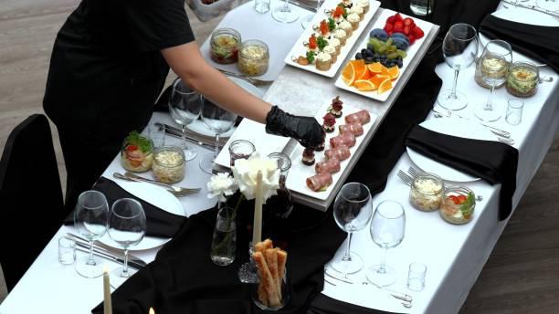 istock Professional waiter preparing elegant appetizers, wearing black gloves on crisp white tablecloth, surrounded by black napkins, empty wine glasses, candlelight 2206564217