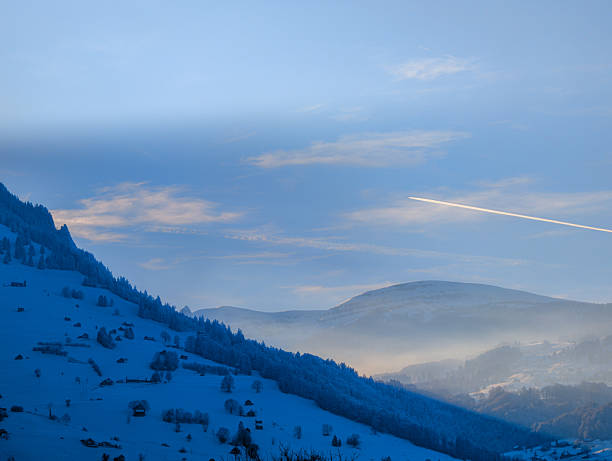 A breathtaking winter landscape featuring snow-covered mountains and a serene sky. A bright airplane contrail cuts across the horizon, adding a dynamic element to the tranquil scene. The soft morning light enhances the misty atmosphere, creating a dreamy winter mood