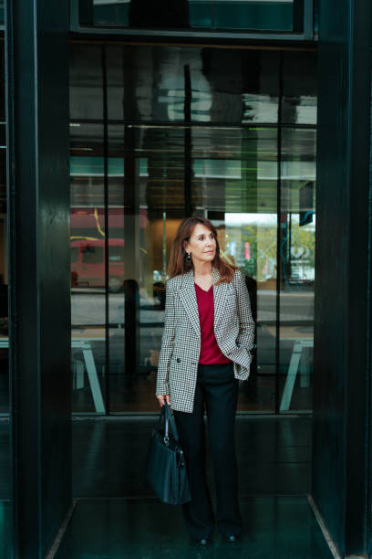 Businesswoman leaving office building with hand in pocket Elegant businesswoman walking out of a modern office building, carrying a black handbag and looking away walking into glass door stock pictures, royalty-free photos & images