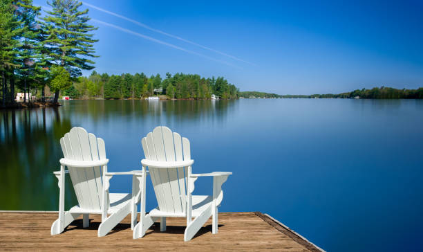 istock White chairs on a dock face a tranquil lake, framed by cottages. 2206582501