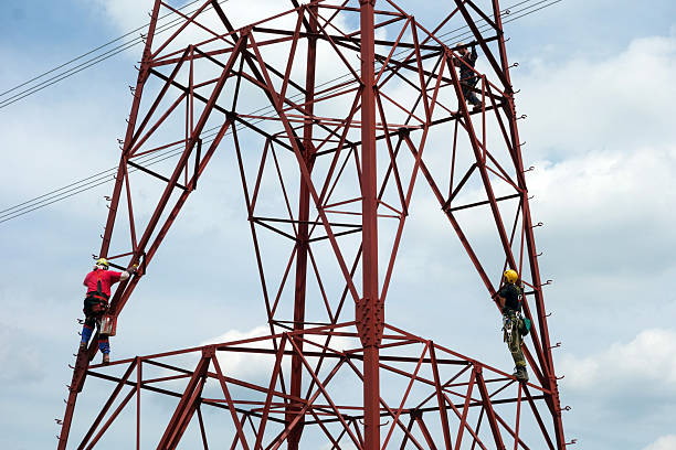 A low-angle shot captures two workers scaling a towering red electrical transmission tower against a pale, overcast sky. The tower's intricate steel lattice structure forms a stark geometric pattern, contrasting with the soft, diffused light. The workers, clad in safety harnesses and work gear, appear as small figures against the massive framework, emphasizing the scale of the structure and the height at which they are working. Thin electrical wires stretch horizontally across the frame, adding to the industrial aesthetic. The image highlights the challenging and potentially dangerous work of maintaining essential infrastructure.