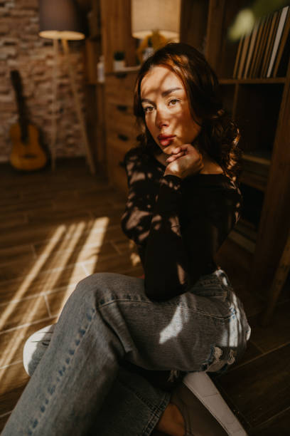 Young woman sitting on the floor in her living room, posing while illuminated by warm sunlight filtering through the window, creating shadows and highlights
