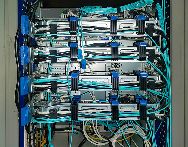 Back side of network servers in the rack mounted on skids with cable connections. Black power cables, vents, cyan fiber optic cables fixed in fasteners