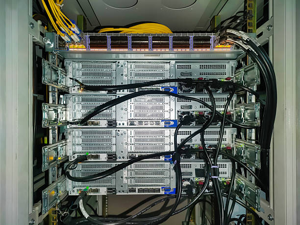 Back side of four network servers in the rack mounted on skids with cable connections and switch on top. Black power cables, vents, yellow fiber optic cables.