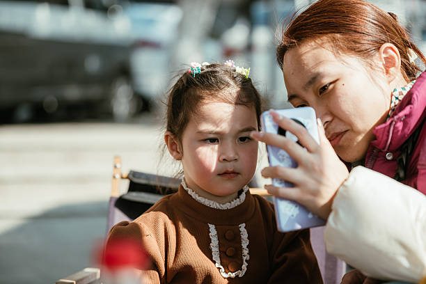 Young Asian mother with her cute multiracial daughter making a photos with mobile phone camera  in cafeteria in the city park during  a rest from a long walk