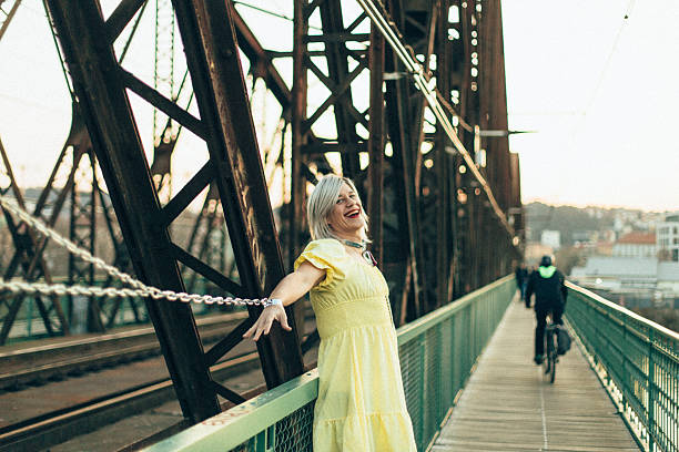 An individual stands on a pedestrian bridge while elegantly dressed in a vibrant yellow dress, with arms outstretched, as a cyclist swiftly passes by during a beautiful sunset