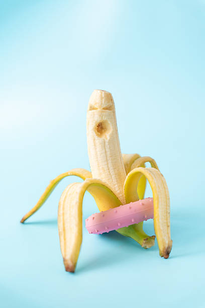 A banana sits upright, partially peeled, with a pink donut resting on its peel, on a light blue background.
