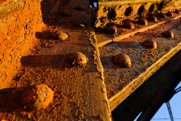 A close-up shot captures the rich textures of a rusted steel truss beam, bathed in warm golden sunlight. The corroded surface and weathered rivets tell a story of time, endurance, and industrial decay. Shadows and highlights emphasize the intricate details, creating a dramatic interplay between light and texture.